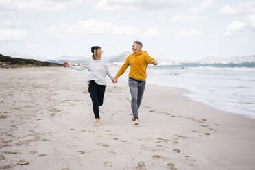 Happy mature couple holding hands and running at beach - JOSEF11405