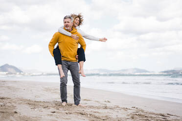 Carefree mature woman enjoying piggyback ride at beach - JOSEF11407