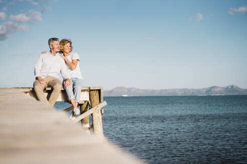 Happy mature couple sitting on jetty over sea in front of sky - JOSEF11426