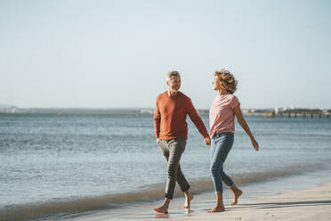 Happy mature couple walking on shore at beach - JOSEF11430