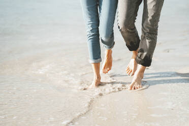 Mature couple walking barefoot on shore at beach - JOSEF11433