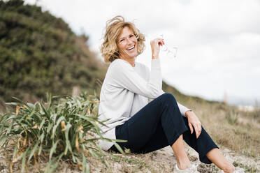 Happy mature woman with eyeglasses sitting by plant at beach - JOSEF11463