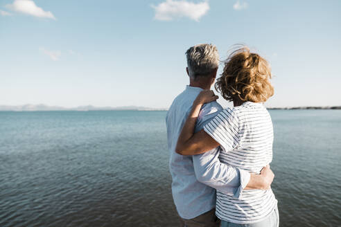 Mature couple with arms around looking at sea on sunny day - JOSEF11503