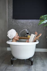 Mature woman reading book in bathtub at home - VEGF05750