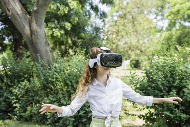 Mädchen mit Virtual-Reality-Simulator steht mit ausgestreckten Armen im Park - OSF00452