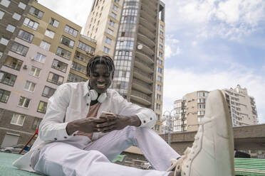 Smiling young man using smart phone sitting in front of buildings - VPIF06853