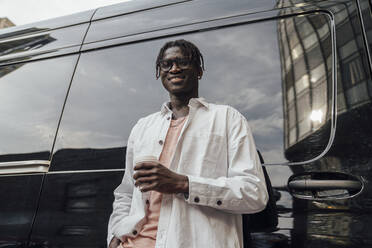 Smiling young man with disposable coffee cup standing in front of van - VPIF06884