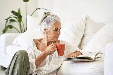 Mature woman with mug reading book in living room - VEGF05794