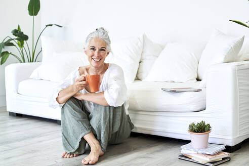 Happy woman with coffee mug sitting in front of sofa at home - VEGF05823