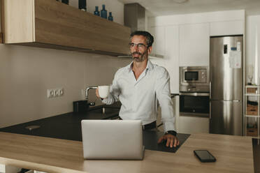 Geschäftsmann mit Kaffeetasse, Smartphone und Laptop am Küchentisch - DMGF00812