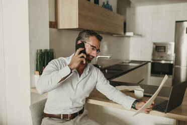 Geschäftsmann, der in der Küche zu Hause ein Dokument liest und mit seinem Smartphone telefoniert - DMGF00814