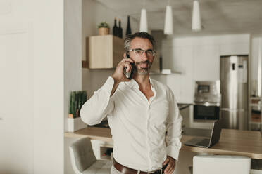 Selbstbewusster Geschäftsmann mit Hand an der Hüfte, der zu Hause in sein Smartphone spricht - DMGF00817