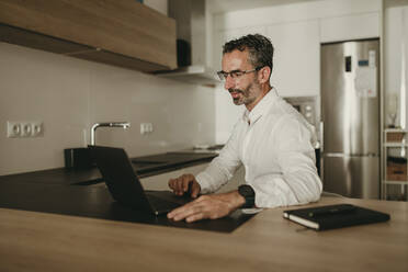 Businessman working on laptop at kitchen counter - DMGF00824