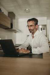 Mature businessman using laptop on kitchen counter at home - DMGF00825
