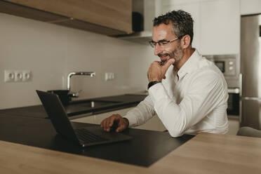 Lächelnder Geschäftsmann mit Hand am Kinn, der zu Hause einen Laptop benutzt - DMGF00826