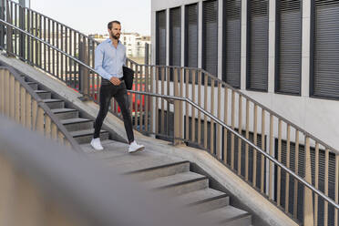 Young businessman with laptop bag walking downwards on staircase - DIGF18308