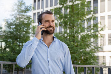 Happy businessman talking on smart phone standing in front of trees - DIGF18319