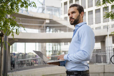 Junger Geschäftsmann mit Laptop steht vor einem Gebäude - DIGF18320