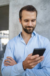 Smiling young businessman using smart phone in front of wall - DIGF18330