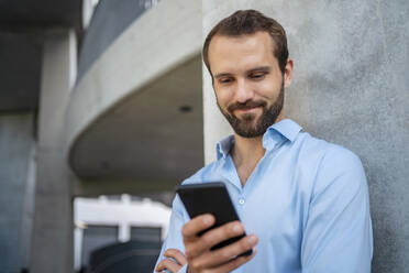 Smiling businessman using mobile phone in front of wall - DIGF18331