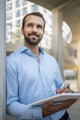 Smiling businessman with tablet leaning on wall - DIGF18344