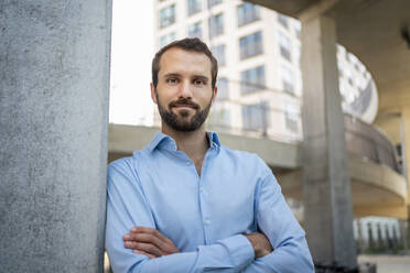 Smiling businessman with arms crossed leaning on wall in front of building - DIGF18346