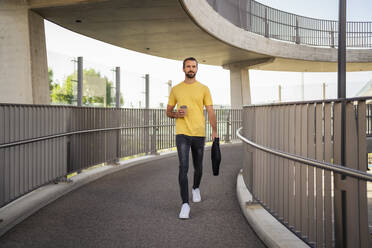 Businessman with disposable coffee cup and bag walking on footbridge - DIGF18352