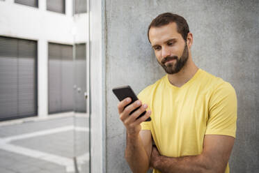 Junger junger Mann mit Bart, der ein Mobiltelefon benutzt und vor einer Wand steht - DIGF18364