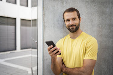 Smiling young man holding mobile phone standing in front of wall - DIGF18365