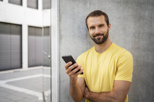 Lächelnder junger Mann mit Mobiltelefon vor einer Wand stehend - DIGF18365