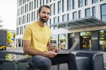 Smiling young freelancer with laptop sitting in front of building - DIGF18370