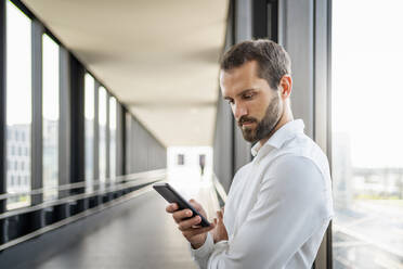 Young businessman using smart phone standing by window on elevated walkway - DIGF18375