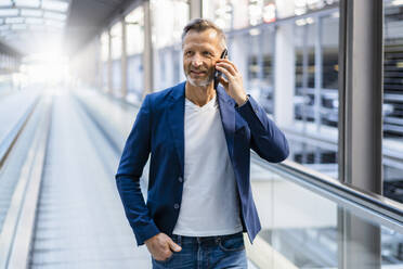 Lächelnder Geschäftsmann mit Hand in der Tasche, der auf einer Rolltreppe mit seinem Smartphone telefoniert - DIGF18394