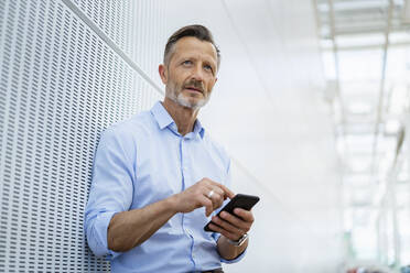 Thoughtful mature businessman with smart phone leaning on wall - DIGF18465