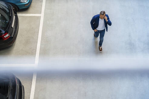 Geschäftsmann, der mit seinem Smartphone telefoniert und an Autos auf einem Parkplatz vorbeiläuft - DIGF18468