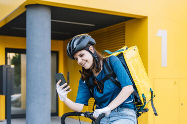 Lächelnde Lieferfrau mit Helm und Smartphone auf dem Fahrrad sitzend - DAMF01039