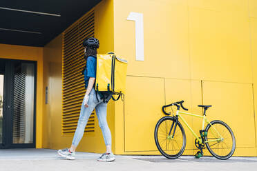 Young delivery woman with backpack walking in front of yellow wall - DAMF01043