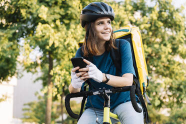 Lächelnde Zustellerin mit Helm und Smartphone auf dem Fahrrad sitzend - DAMF01057