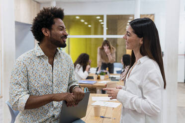 Businesswoman and businessman talking in office with colleagues in background - PNAF04148