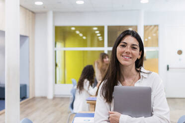 Porträt einer lächelnden Geschäftsfrau, die einen Laptop im Büro hält, mit Kollegen im Hintergrund - PNAF04153