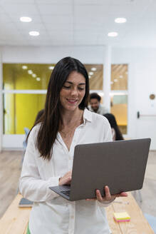 Geschäftsfrau mit Laptop im Büro mit Kollegen im Hintergrund - PNAF04154