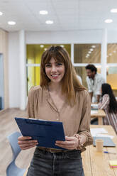 Porträt einer lächelnden Geschäftsfrau, die ein Klemmbrett im Büro hält, mit Kollegen im Hintergrund - PNAF04160