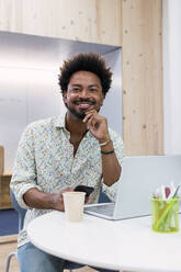 Portrait of smiling businessman with mobile phone and laptop in office - PNAF04276