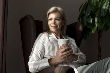 Smiling mature woman with coffee mug sitting in armchair at home - LLUF00732