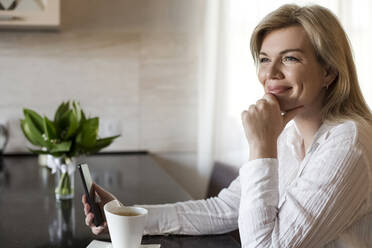Lächelnde Frau mit Smartphone beim Kaffee trinken zu Hause - LLUF00753