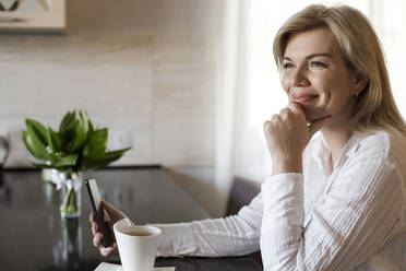 Smiling woman with smart phone having coffee at home - LLUF00753