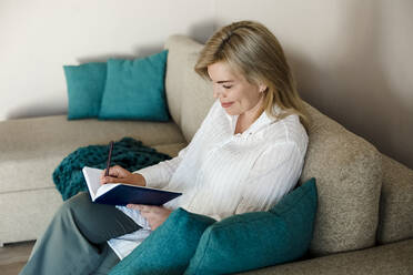 Smiling mature woman writing in diary sitting on couch at home - LLUF00755