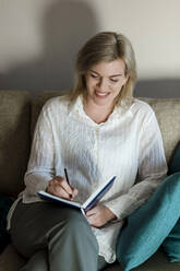 Happy mature woman writing in diary at home - LLUF00756