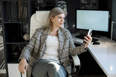 Ältere Geschäftsfrau mit Smartphone sitzt am Computer am Schreibtisch - LLUF00759
