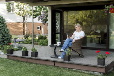 Lächelnde Frau, die ein Smartphone benutzt und auf einem Stuhl auf der Veranda sitzt - LLUF00762